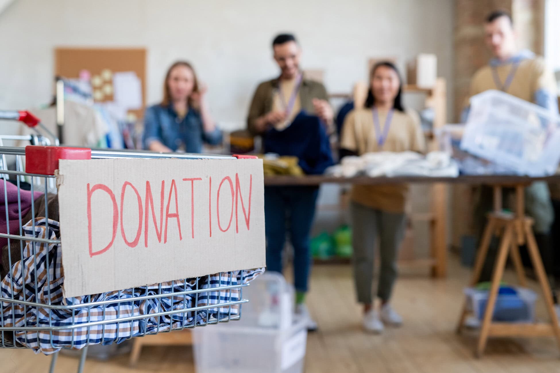 Shopping cart with donation clothes against row of volunteers standing by table Cart with a cardboard sign reading DONATION in red letters, donation items inside; volunteers in the background organizing supplies in a bright room.