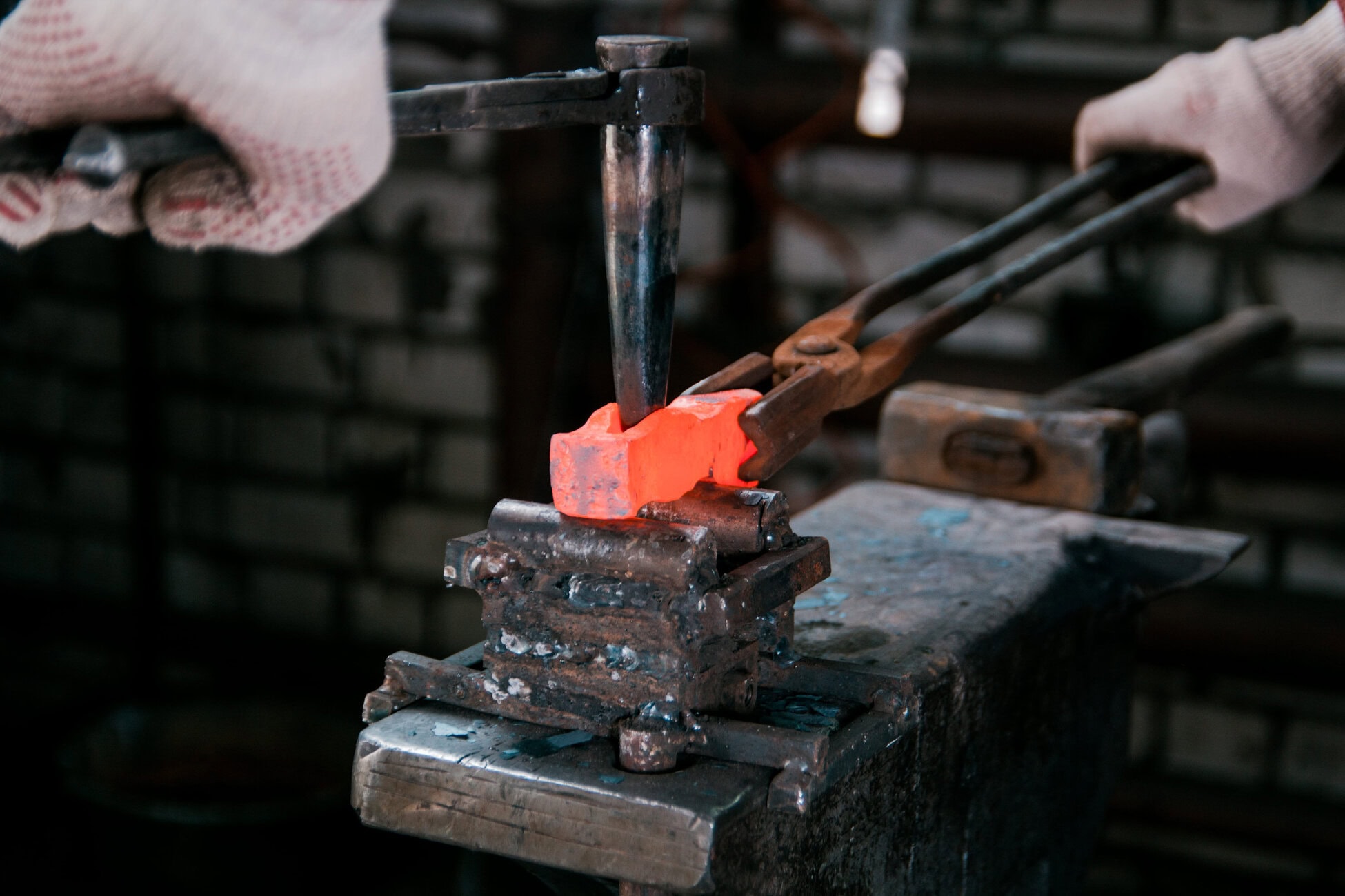 775779 Metric4 Blacksmith wearing gloves shaping a glowing orange-hot metal with tongs and a hammer over an anvil/vise.
