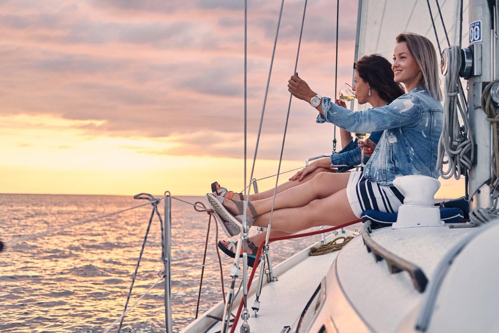 775800 – Ola120 Three women sit on the edge of a sailboat at sunset, sipping wine and enjoying the ocean view.