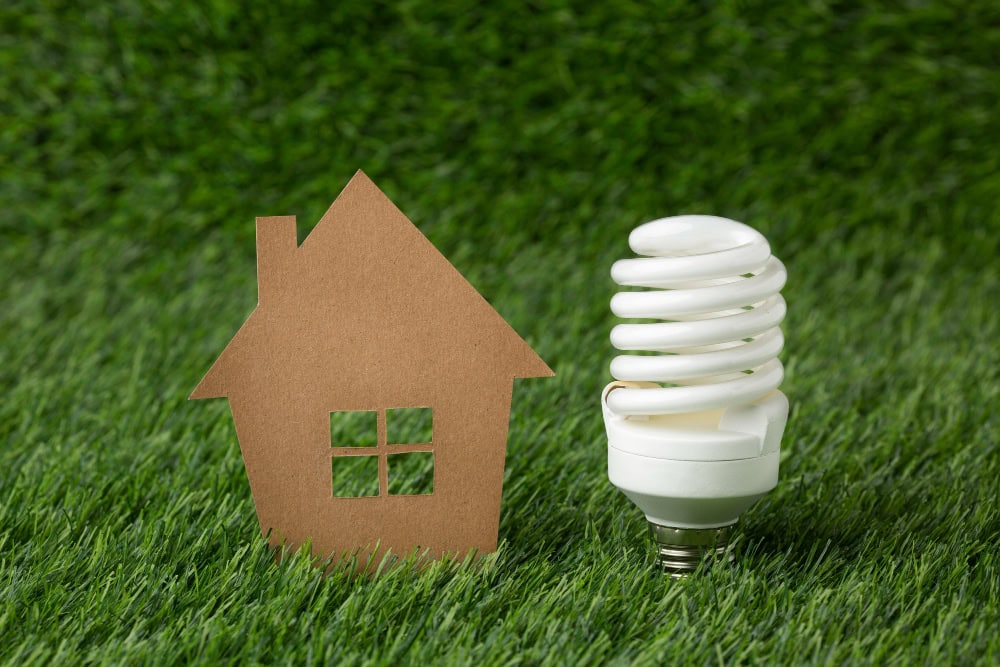 775809 – Electricitat i Aplicacions Germán SL Eco-friendly home energy concept: paper house model beside a compact fluorescent lamp on grass