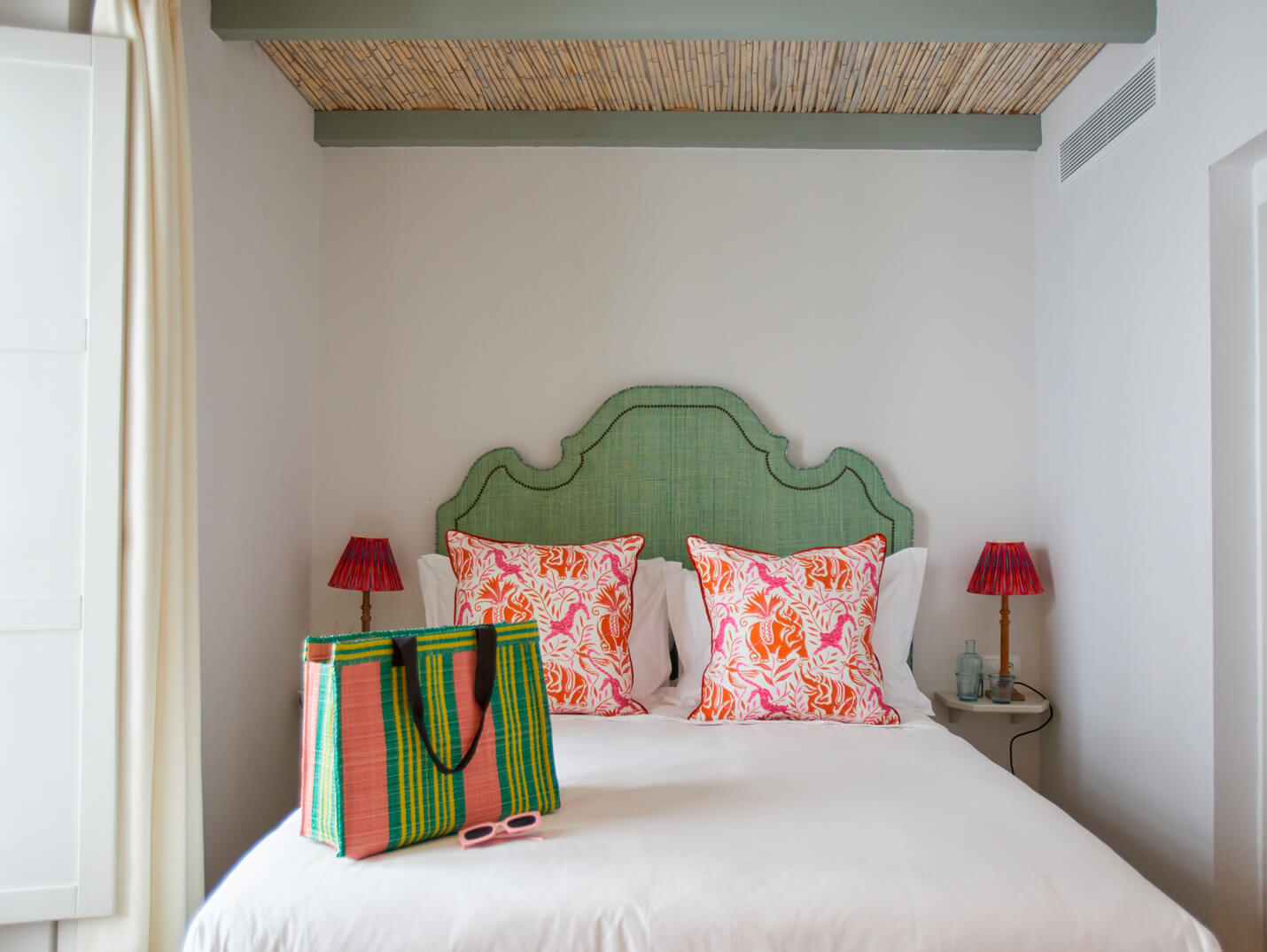 El Escondite – IP Bedroom with a mint-green upholstered headboard, white bedding, and two pink/orange tropical-pattern cushions; a colorful striped tote and pink sunglasses sit on the duvet, with red bedside lamps on either side.