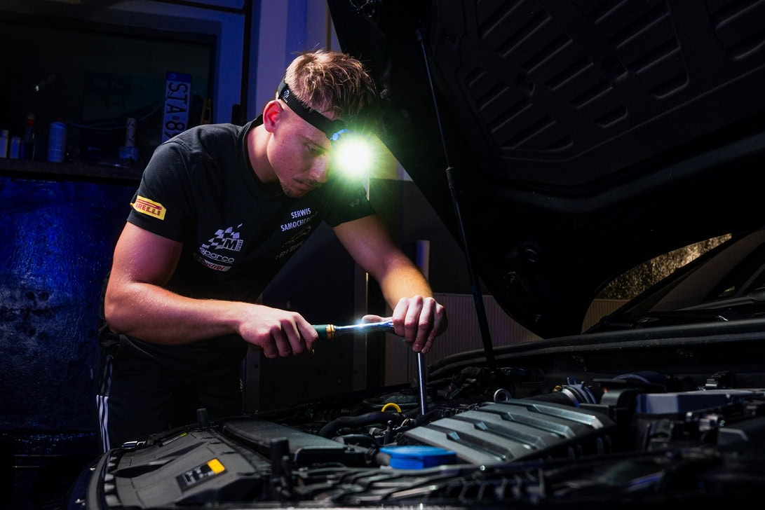 Linterna frontal – Mtech – IP Mechanic with a headlamp inspecting a car engine under the open hood, tool in hand