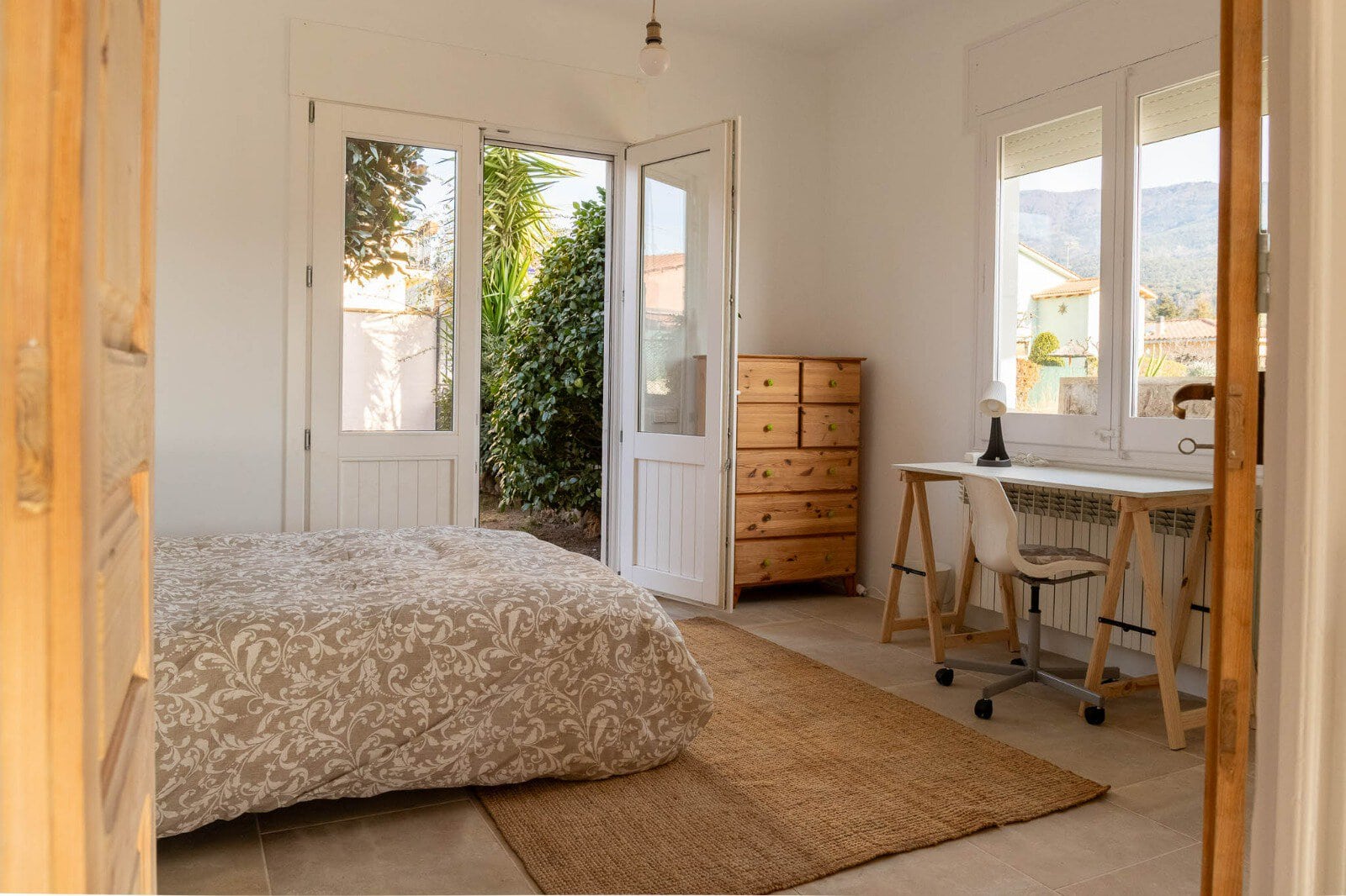 Montseny coliving – IP Bright bedroom with a patterned duvet, open glass doors to a leafy garden, and a wooden dresser.