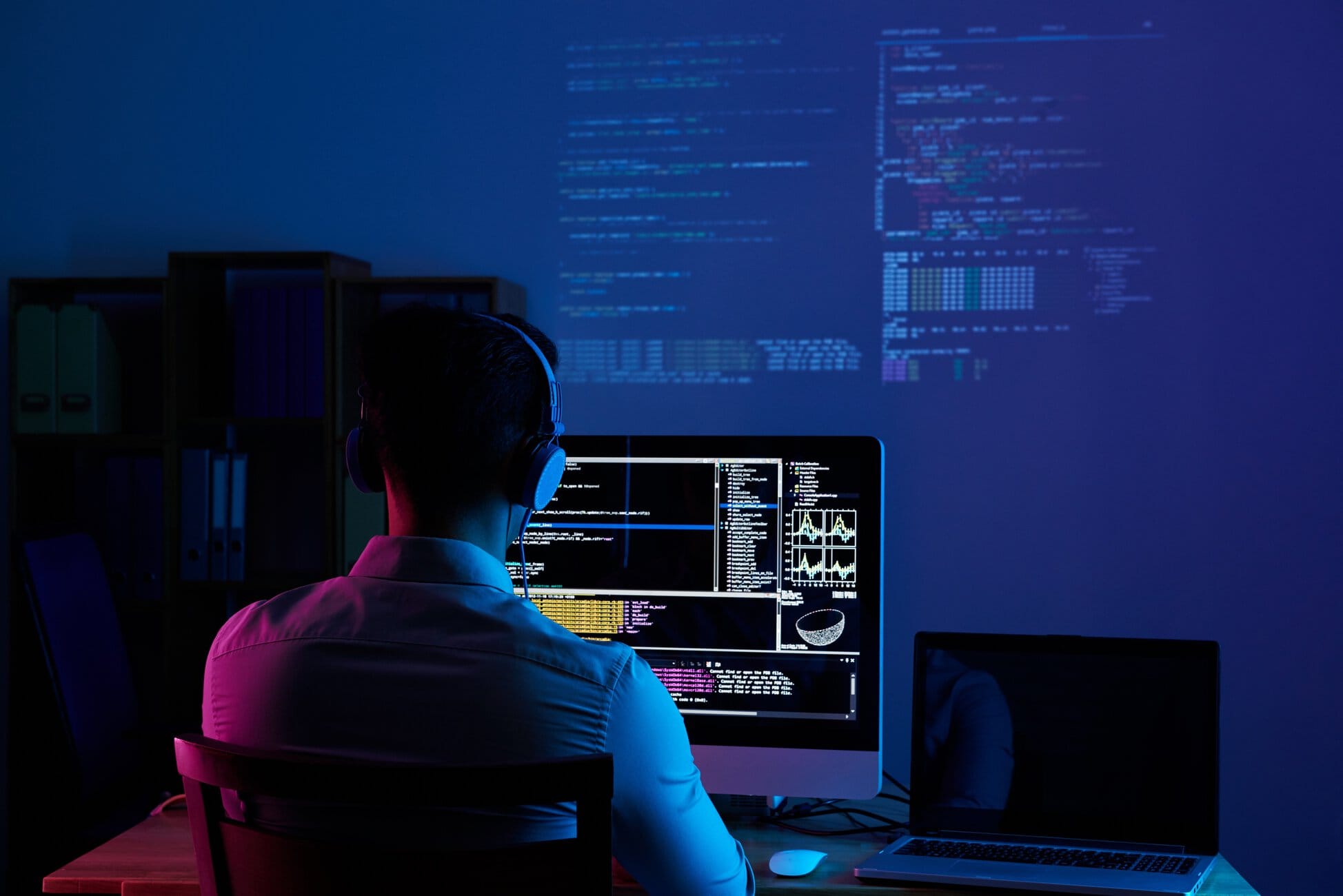 777904 – Data Pro Person with headphones seated at a desk coding on two monitor displays in a dim blue-lit room, with code projected on the wall behind.