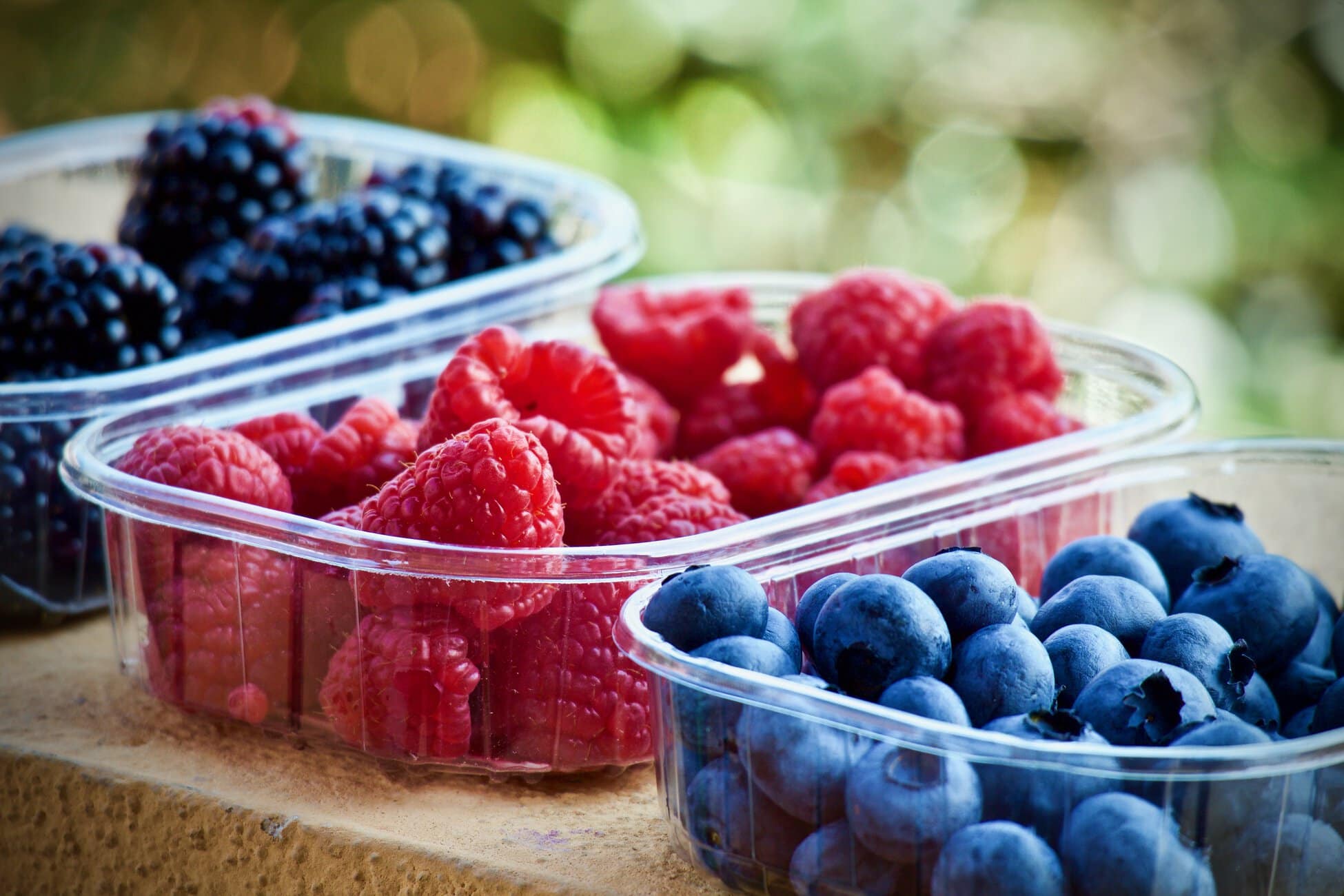 Plastic cups hold fresh raspberries, blueberries, and blackberries on a wooden surface outdoors.