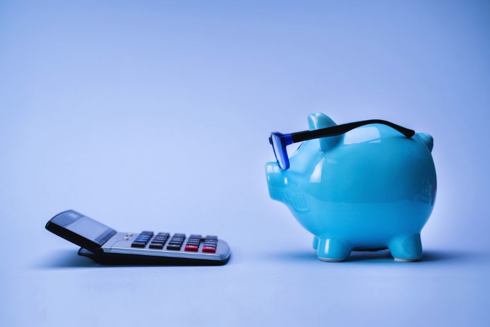 Blue piggy bank wearing glasses sits next to a calculator on a blue background, illustrating savings or budgeting for financial planning.