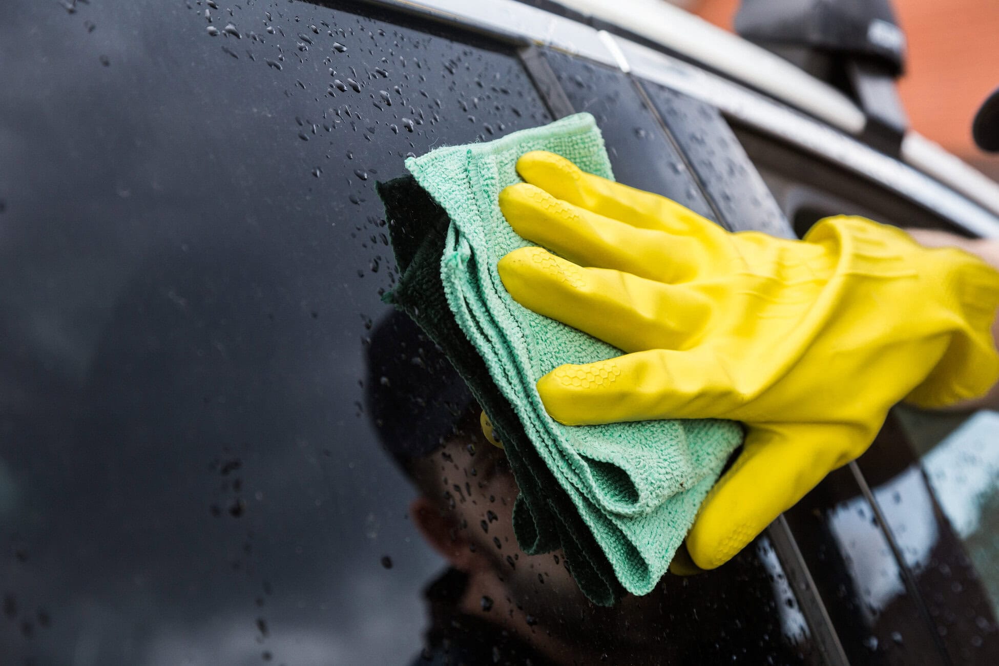 778201 Detailcar Hands in yellow gloves wiping a wet car window with a green microfiber cloth, close-up.
