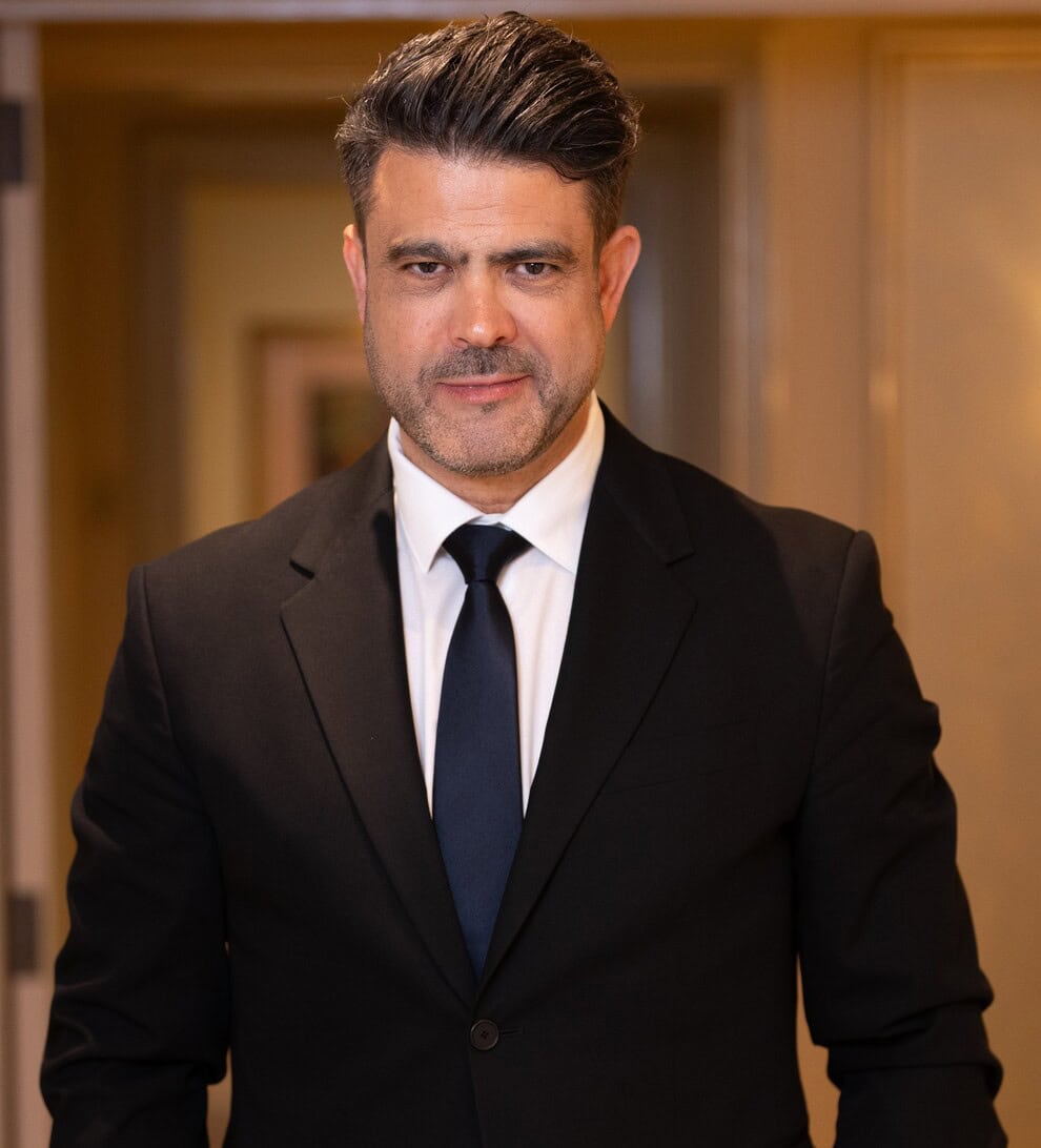 MH_PHOTOGRAPHY Professional portrait: man in a black suit and navy tie standing in a hallway, looking at the camera.