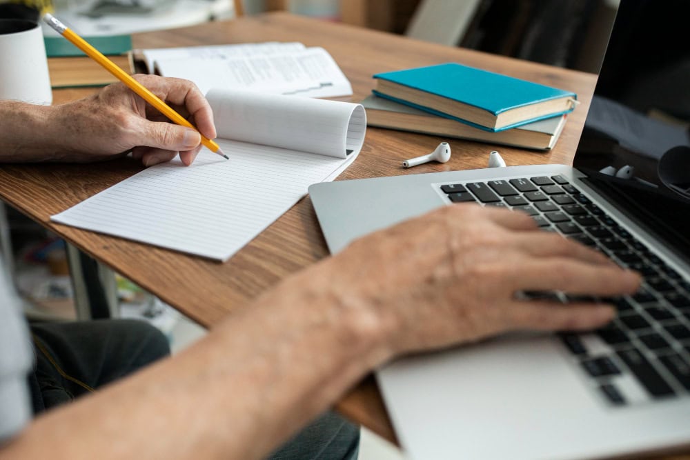 Person writing in a lined notebook at a wooden desk with a laptop, books, and earbuds nearby.