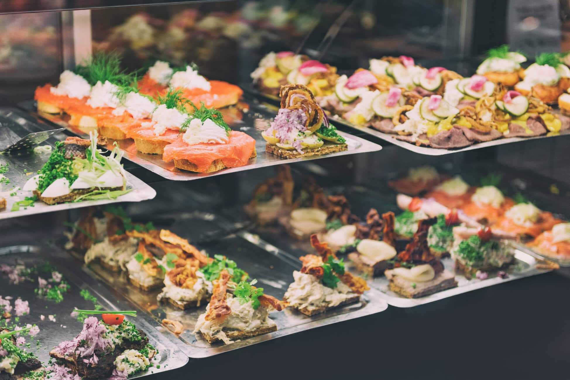 Assorted savory canapés and crostini on metal trays inside a bakery display case, garnished with herbs and vegetables.