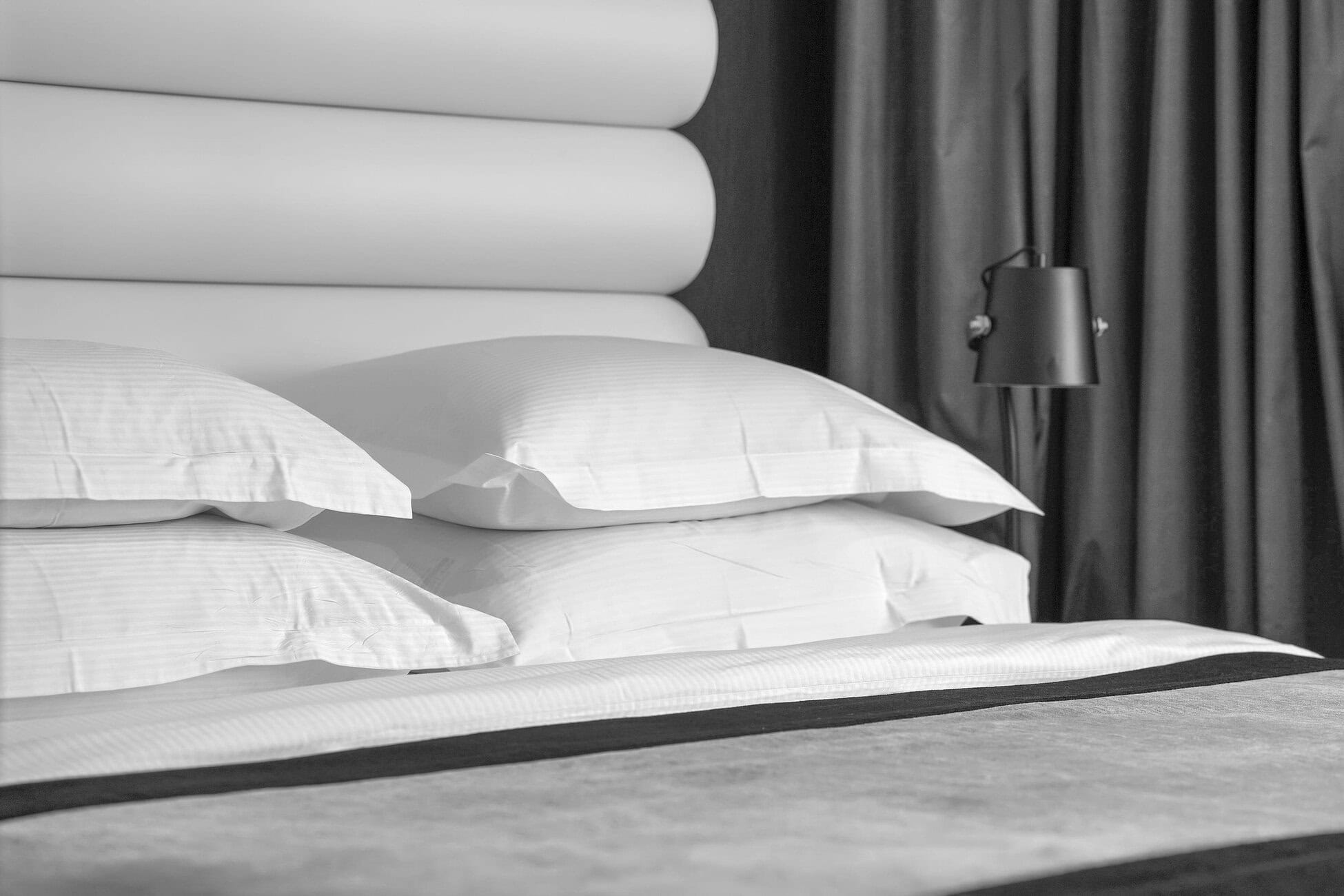 White bed with layered cylindrical headboard cushions, several pillows on the mattress, a black bedside lamp to the right, and dark curtains in the background (black-and-white photo).