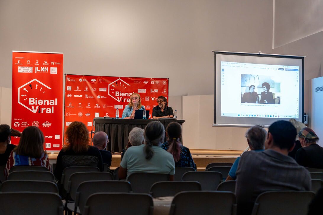 Panel discussion on stage at Bienal Viral with two speakers at a table, red Bienal Viral banners, and an audience watching.