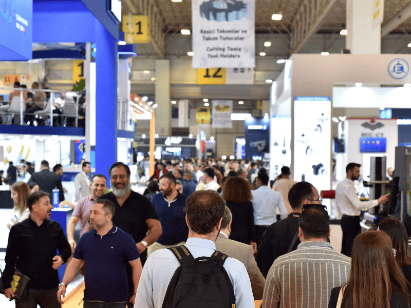 Crowded trade show floor with attendees walking past blue booths and overhead banners.