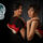Two women stand back-to-back in black outfits, each holding red sneakers, against a dark backdrop with a teal and white TRAN logo to the left.